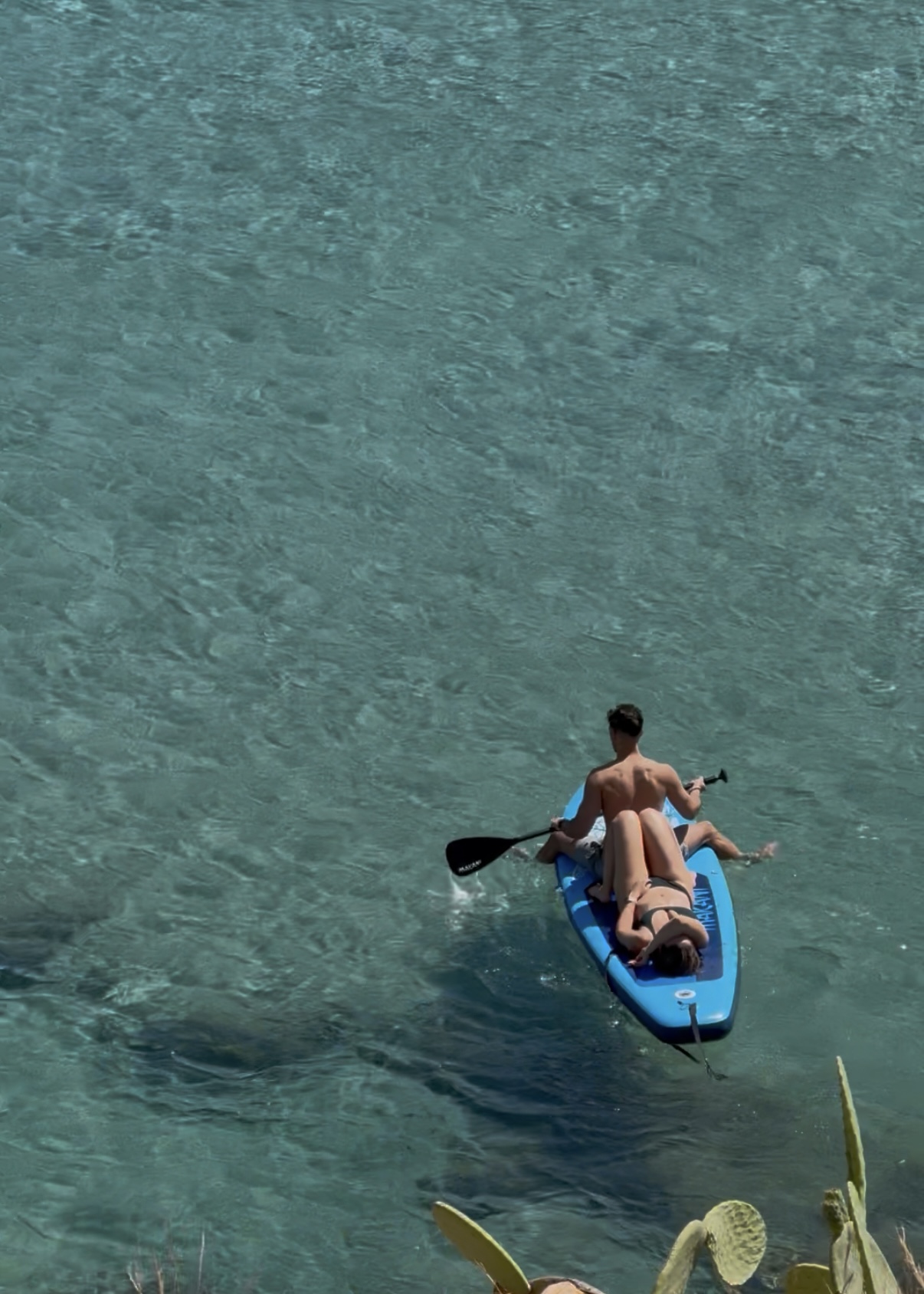 A man and woman share a waterboard - she is lying down while he is paddling them forward over crystal blue water.